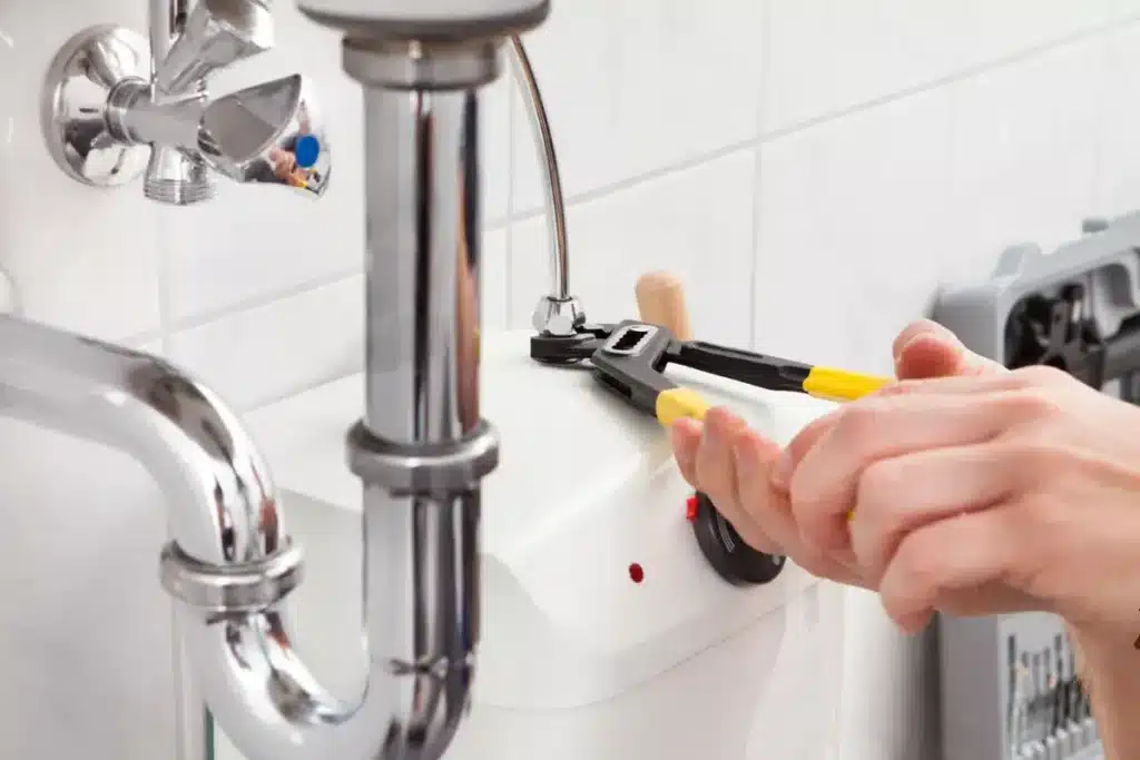 Plombier utilisant une pince pour serrer le raccord d'un petit chauffe-eau mural blanc, près d'un siphon et d'une vanne d'arrêt chrome.